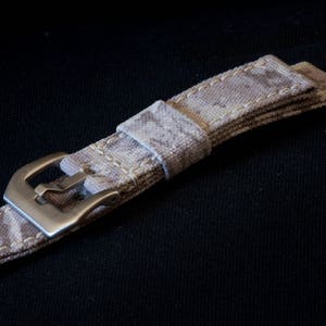 May include: A beige canvas watch strap with a silver buckle. The strap has a subtle pattern and is stitched with white thread.