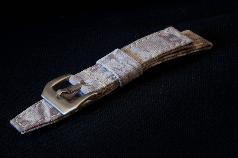 May include: A beige canvas watch strap with a silver buckle. The strap has a subtle pattern and is stitched with white thread.