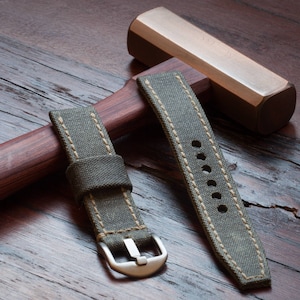 May include: Two olive green canvas watch straps with beige stitching and a silver buckle. The straps are displayed on a wooden surface with a wooden hammer in the background. The watch straps are new.