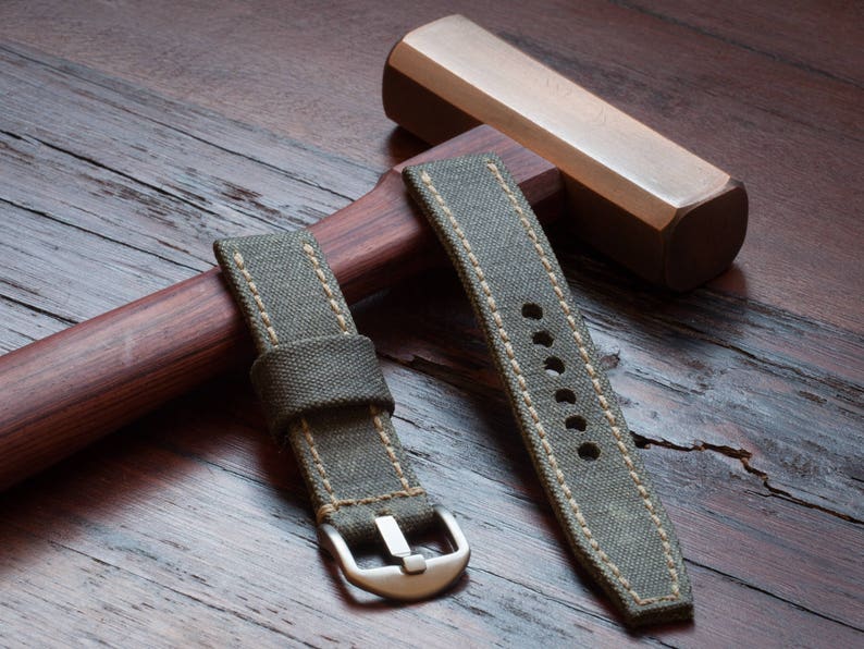 May include: Two olive green canvas watch straps with beige stitching and a silver buckle. The straps are displayed on a wooden surface with a wooden hammer in the background. The watch straps are new.