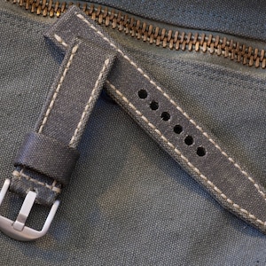 May include: A gray canvas watch strap with white stitching and a silver buckle. The strap has multiple adjustment holes and is set against a textured background with a zipper.
