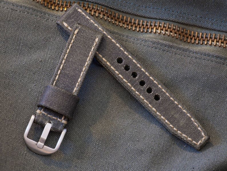 May include: A gray canvas watch strap with white stitching and a silver buckle. The strap has multiple adjustment holes and is set against a textured background with a zipper.