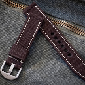 May include: A brown canvas watch strap with white stitching and a silver buckle. The strap has a series of holes for adjusting the fit.