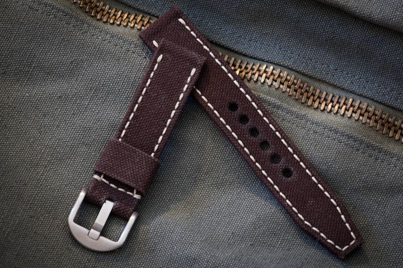 May include: A brown canvas watch strap with white stitching and a silver buckle. The strap has a series of holes for adjusting the fit.