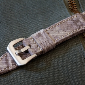 May include: A close-up of a beige and brown patterned watch strap with a silver buckle. The strap is made of fabric and has a stitched edge.