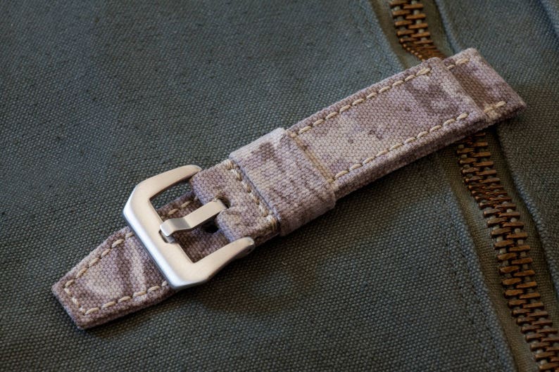 May include: A close-up of a beige and brown patterned watch strap with a silver buckle. The strap is made of fabric and has a stitched edge.