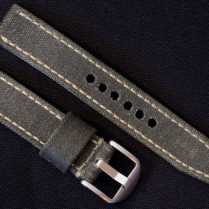 May include: A gray canvas watch strap with off-white stitching and a silver buckle. The strap has multiple holes for adjustment and is displayed on a dark blue textured surface.