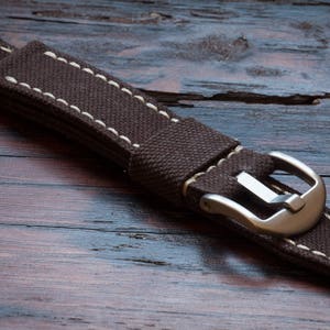 May include: A brown canvas watch strap with white stitching and a silver buckle.