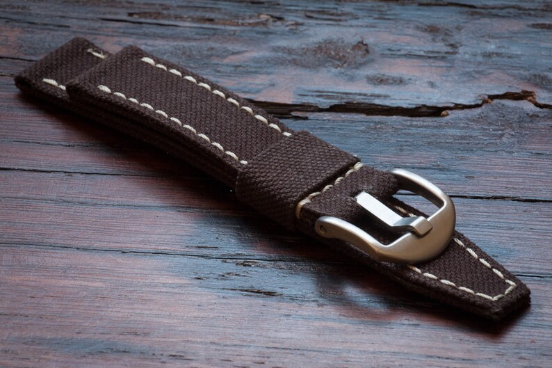 May include: A brown canvas watch strap with white stitching and a silver buckle.