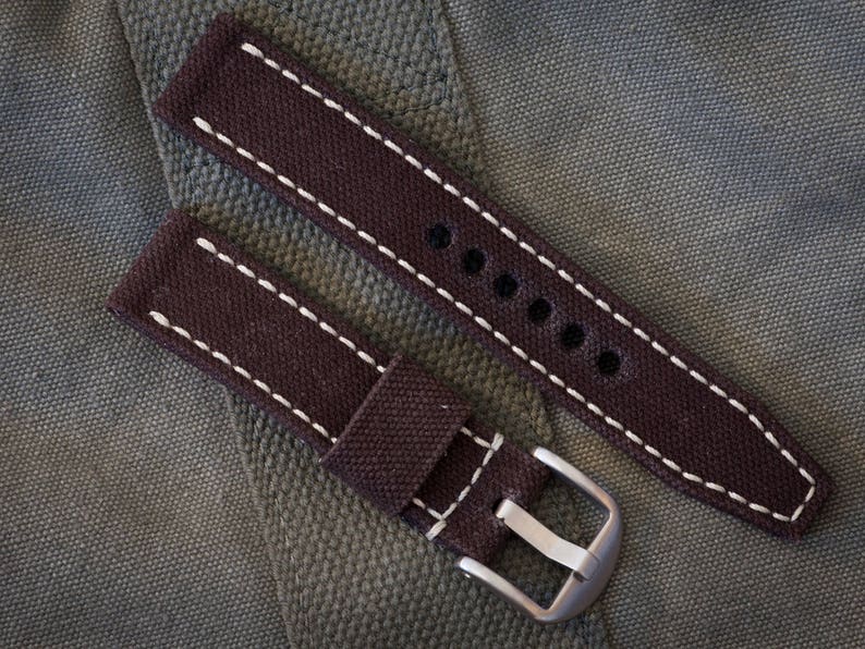 May include: Two brown canvas watch straps with white stitching. The straps have metal buckles and are designed for a wristwatch.