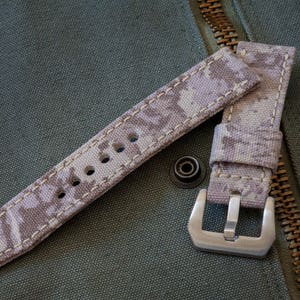 May include: A beige watch strap with a camouflage pattern and white stitching. The strap has a silver buckle.