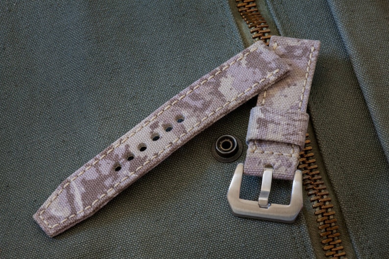 May include: A beige watch strap with a camouflage pattern and white stitching. The strap has a silver buckle.