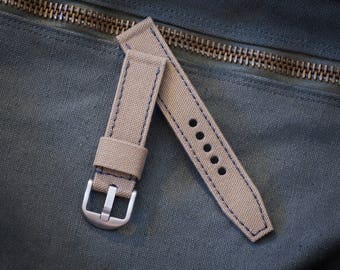 May include: A tan canvas watch strap with blue stitching and a silver buckle. The strap is lying on a dark gray fabric surface.