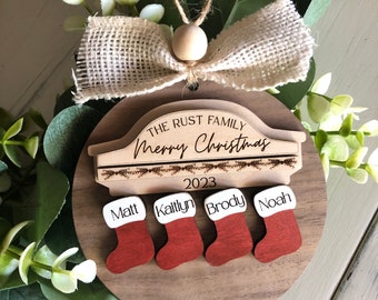 May include: A wooden Christmas ornament with a car silhouette and the text "The Rust Family Merry Christmas 2023". Four red stockings with white trim are attached to the ornament, each with a name: Matt, Kaitlyn, Brody, and Noah.
