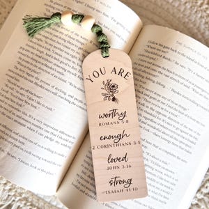 May include: Wooden bookmark with a green tassel and wooden beads. The bookmark has the words "YOU ARE" at the top, followed by "worthy, enough, loved, strong" and Bible verses. The bookmark is resting on the pages of a book.