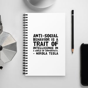 May include: A white spiral bound notebook with a black quote on the front that reads "Anti-social behavior is a trait of intelligence in a world of conformists. - Nikola Tesla". The notebook is on a white surface with a coffee cup, a pencil, and a black smartphone.