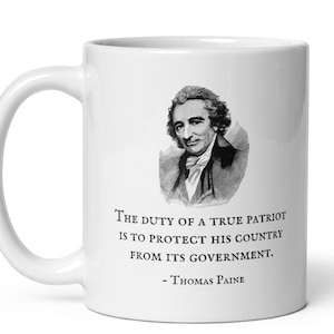 K&ouml;nnte beinhalten: Wei&szlig;e Keramiktasse mit einem Schwarz-Wei&szlig;-Portr&auml;t von Thomas Paine und einem Zitat. Das Zitat lautet: "Die Pflicht eines wahren Patrioten ist es, sein Land vor seiner Regierung zu sch&uuml;tzen."