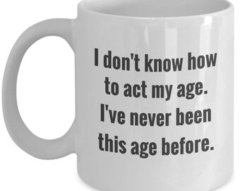 May include: White ceramic coffee mug with black text that reads "I don't know how to act my age. I've never been this age before."