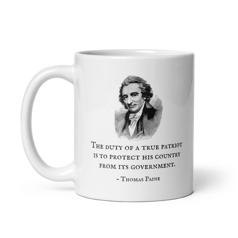 K&ouml;nnte beinhalten: Wei&szlig;e Keramiktasse mit einem Schwarz-Wei&szlig;-Portr&auml;t von Thomas Paine und einem Zitat. Das Zitat lautet: "Die Pflicht eines wahren Patrioten ist es, sein Land vor seiner Regierung zu sch&uuml;tzen."