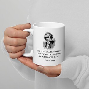 K&ouml;nnte beinhalten: Wei&szlig;e Keramik-Tasse mit einem Portr&auml;t von Thomas Paine und dem Zitat: &bdquo;Die Pflicht eines wahren Patrioten ist es, sein Land vor seiner Regierung zu sch&uuml;tzen.&ldquo; Die Tasse hat einen gebogenen Griff.
