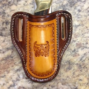 May include: A brown leather knife sheath with a decorative stitching pattern and a tooled design of a bull's head. The sheath has a belt loop and a snap closure.