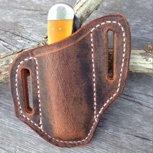 May include: A brown leather knife sheath with white stitching. The sheath has two belt loops and is designed to hold a pocket knife.
