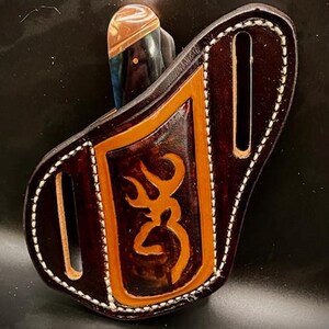 May include: A brown leather knife sheath with white stitching detail. The sheath has a buck logo embossed on the front.
