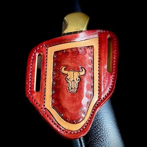 May include: Red leather knife sheath with a tan leather trim and a tooled design of a bull skull.