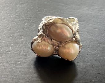 May include: A silver ring featuring three large, lustrous pearls. The ring has a textured, organic design, with the pearls set in a cluster. The pearls have a creamy, off-white color, and the ring appears to be handmade.