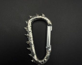 May include: A silver-toned carabiner featuring a textured, irregular surface with multiple pointed, thorn-like protrusions along the outer edge. The carabiner includes a smooth, straight spring-loaded gate mechanism on the right side for opening.