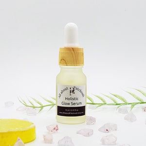 Peut inclure: Petite bouteille en verre transparent de s&eacute;rum Holistic Glow, avec un compte-gouttes blanc et un bouchon couleur bambou. L'&eacute;tiquette indique le nom de la marque et les informations sur le produit. Le fond comprend des cristaux d&eacute;coratifs et une tranche de citron.