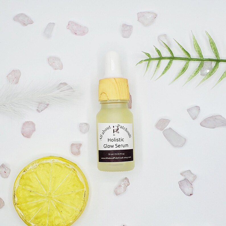Peut inclure: Petite bouteille en verre de "Holistic Glow Serum" avec un bouchon et une pipette jaunes. L'&eacute;tiquette indique "All about Patchouli" et "10 ml / 0.33 fl oz." La bouteille est entour&eacute;e d'&eacute;l&eacute;ments d&eacute;coratifs, dont une tranche de citron, des cristaux et une foug&egrave;re.