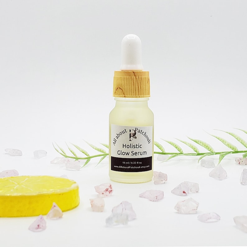 Peut inclure: Petite bouteille en verre transparent de s&eacute;rum Holistic Glow, avec un compte-gouttes blanc et un bouchon couleur bambou. L'&eacute;tiquette indique le nom de la marque et les informations sur le produit. Le fond comprend des cristaux d&eacute;coratifs et une tranche de citron.