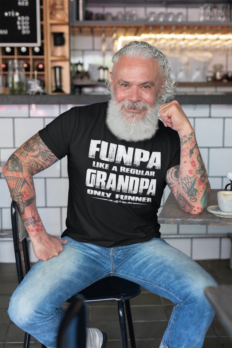 May include: A man wearing a black t-shirt with the text "Funpa like a regular Grandpa only funnier" in white lettering. He is sitting on a chair with his arms resting on a table.