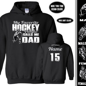 May include: Black hooded sweatshirt with a white graphic that says "My Favourite Hockey Player Calls Me Dad". The graphic includes a hockey player with a stick and puck. The back of the sweatshirt has a white graphic with the word "Name" and the number "15".