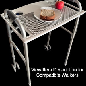 May include: A white walker tray with a gray non-slip mat, a cup holder, and handles. The tray is placed on top of a silver walker with wheels. A red apple, a slice of bread on a white plate, and a clear water bottle are on the tray.