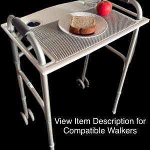 May include: A silver walker tray with a textured surface, a cup holder, and handles. A plate with a slice of bread, a red apple, and a water bottle are on the tray. The text "View Item Description for Compatible Walkers" is visible.