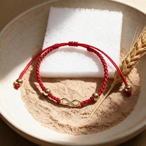 Puede incluir: Una pulsera de cordón trenzado rojo con un dije de símbolo de infinito en tono dorado incrustado con pequeñas piedras claras. La pulsera incluye dos cuentas esféricas doradas pulidas y extremos ajustables con cuentas doradas.
