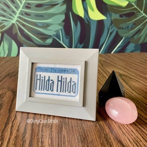 May include: A framed cross-stitch with the text "Hilda Hilda" in blue thread on a white background. The frame is light gray and has a white mat. The cross-stitch is sitting on a wooden surface with a black and a pink crystal.