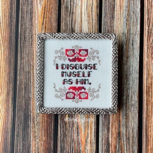 May include: A framed cross-stitch with a white background and red and gold embroidery. The text reads "I disguise myself as him."