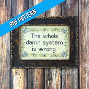 May include: A cross-stitch embroidery framed in a dark brown wood frame. The embroidery is on a cream-colored fabric with green and blue floral accents. The text reads "The whole darn system is wrong."