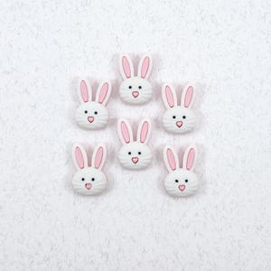 May include: Seven white bunny face embellishments with pink ears and heart-shaped noses are arranged on a textured white surface. The bunny faces have black dot eyes and are suitable for crafts.