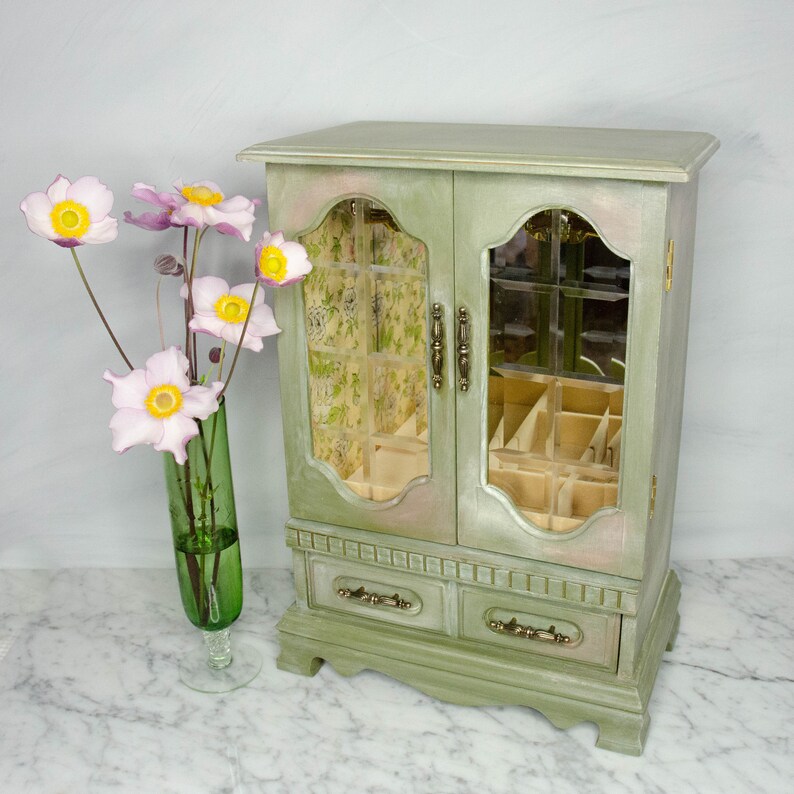 K&ouml;nnte beinhalten: Eine gr&uuml;ne Holzk&auml;stchen f&uuml;r Schmuck mit zwei T&uuml;ren und zwei Schubladen. Die T&uuml;ren haben Glasscheiben mit einem Blumenmuster. Die Schachtel steht auf einer wei&szlig;en Marmorfl&auml;che.