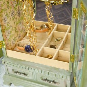 K&ouml;nnte beinhalten: Eine gr&uuml;ne Holzk&auml;stchen f&uuml;r Schmuck mit einem verspiegelten Innenraum und mehreren F&auml;chern. Die Box hat ein Blumenmuster im Inneren und eine goldene Kette, die von oben h&auml;ngt.