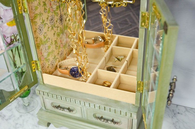 K&ouml;nnte beinhalten: Eine gr&uuml;ne Holzk&auml;stchen f&uuml;r Schmuck mit einem verspiegelten Innenraum und mehreren F&auml;chern. Die Box hat ein Blumenmuster im Inneren und eine goldene Kette, die von oben h&auml;ngt.