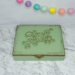 May include: A green wooden box with a decorative design of gold metal circles and a gold metal latch. The box is on a white marble surface.