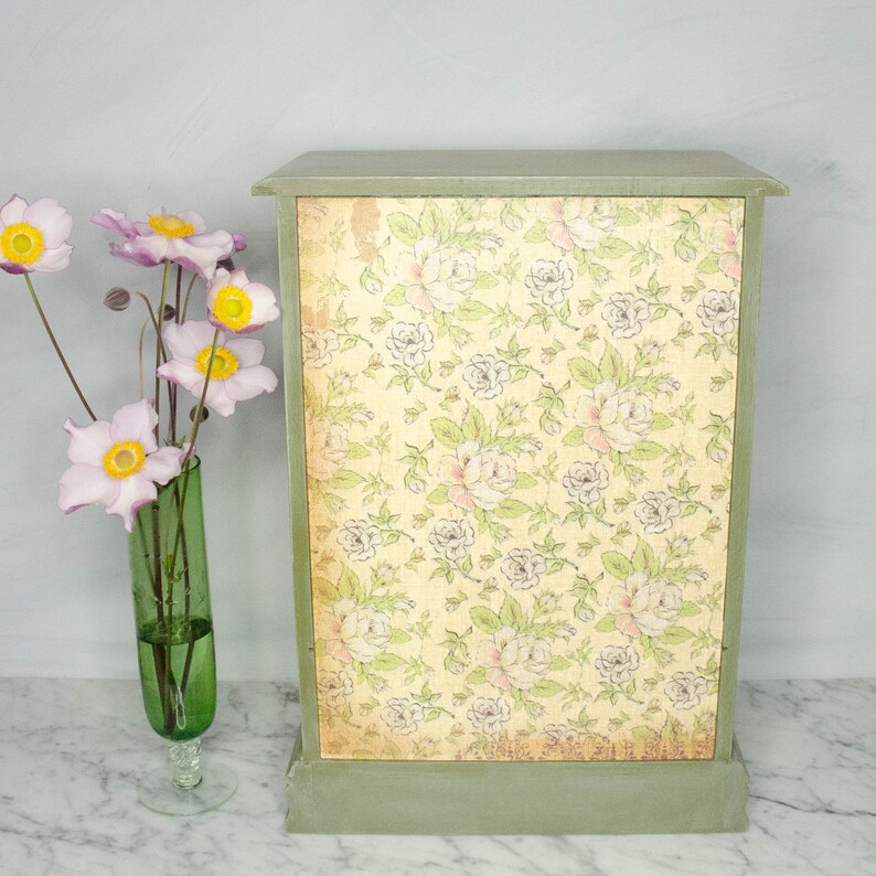 K&ouml;nnte beinhalten: Ein gr&uuml;ner Holzschrank mit einem Blumendesign auf der Vorderseite. Der Schrank hat eine einzelne T&uuml;r und einen Sockel.