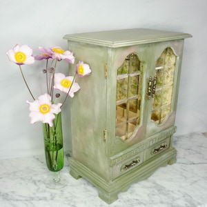 K&ouml;nnte beinhalten: Eine gr&uuml;ne Holzk&auml;stchen f&uuml;r Schmuck mit zwei Glast&uuml;ren und zwei Schubladen. Die Box hat eine abgenutzte Lackierung und ist mit Blumenmustern verziert. Die Box steht auf einer wei&szlig;en Marmorfl&auml;che.