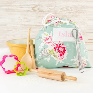 May include: A floral print fabric bag with a pink label that says "Isla". The bag is filled with baking supplies, including a wooden rolling pin, a wooden spoon, a whisk, and a cookie cutter.