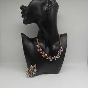 May include: A gold necklace and brooch set with iridescent pink and blue leaves. The necklace is on a black mannequin bust.